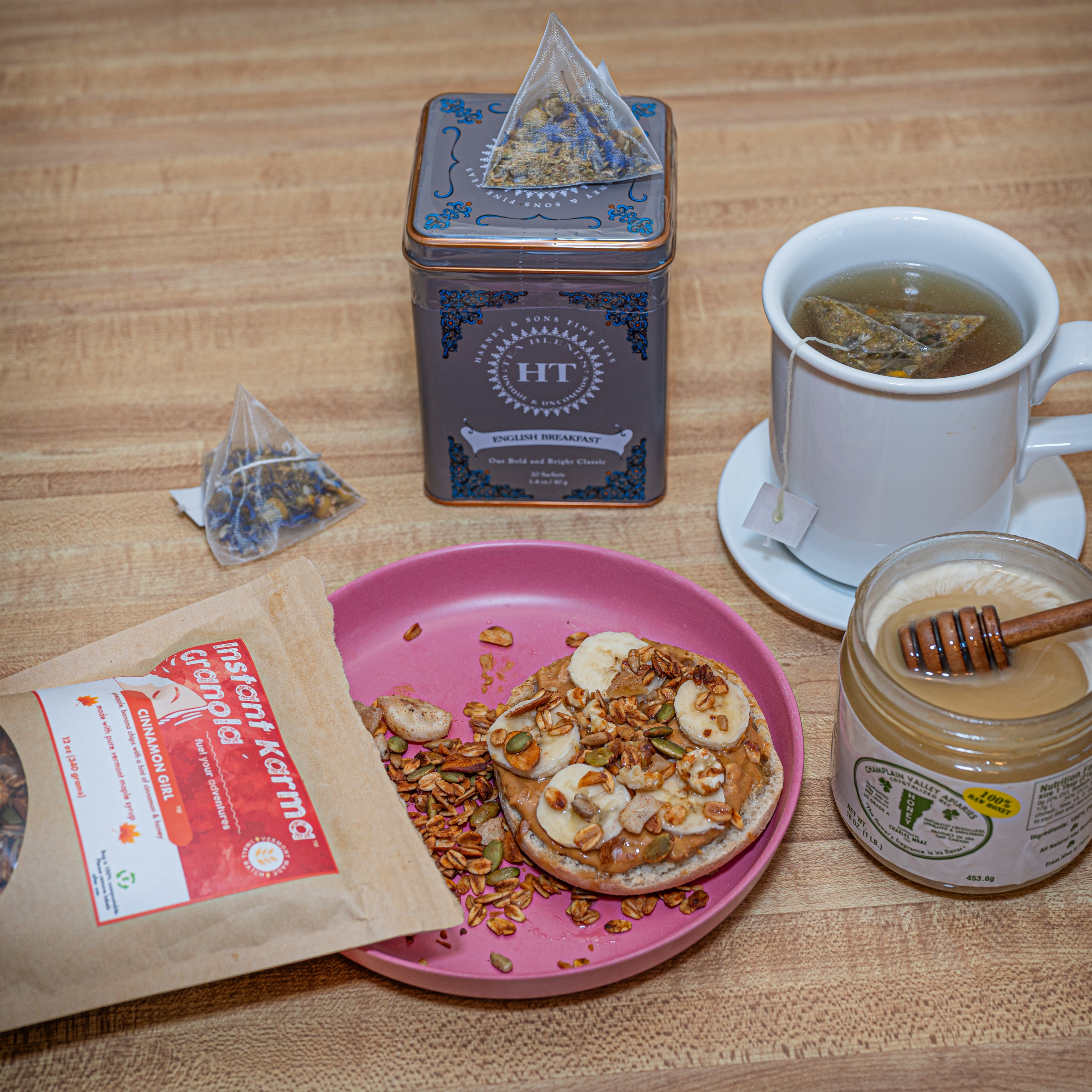 Tableau of mug of steeped tea, jar of Champlain Valley Honey, tin of Harney & Sons English Breakfast tea and plate with toast topped with peanut butter, sliced bananas sprinkled with granola
