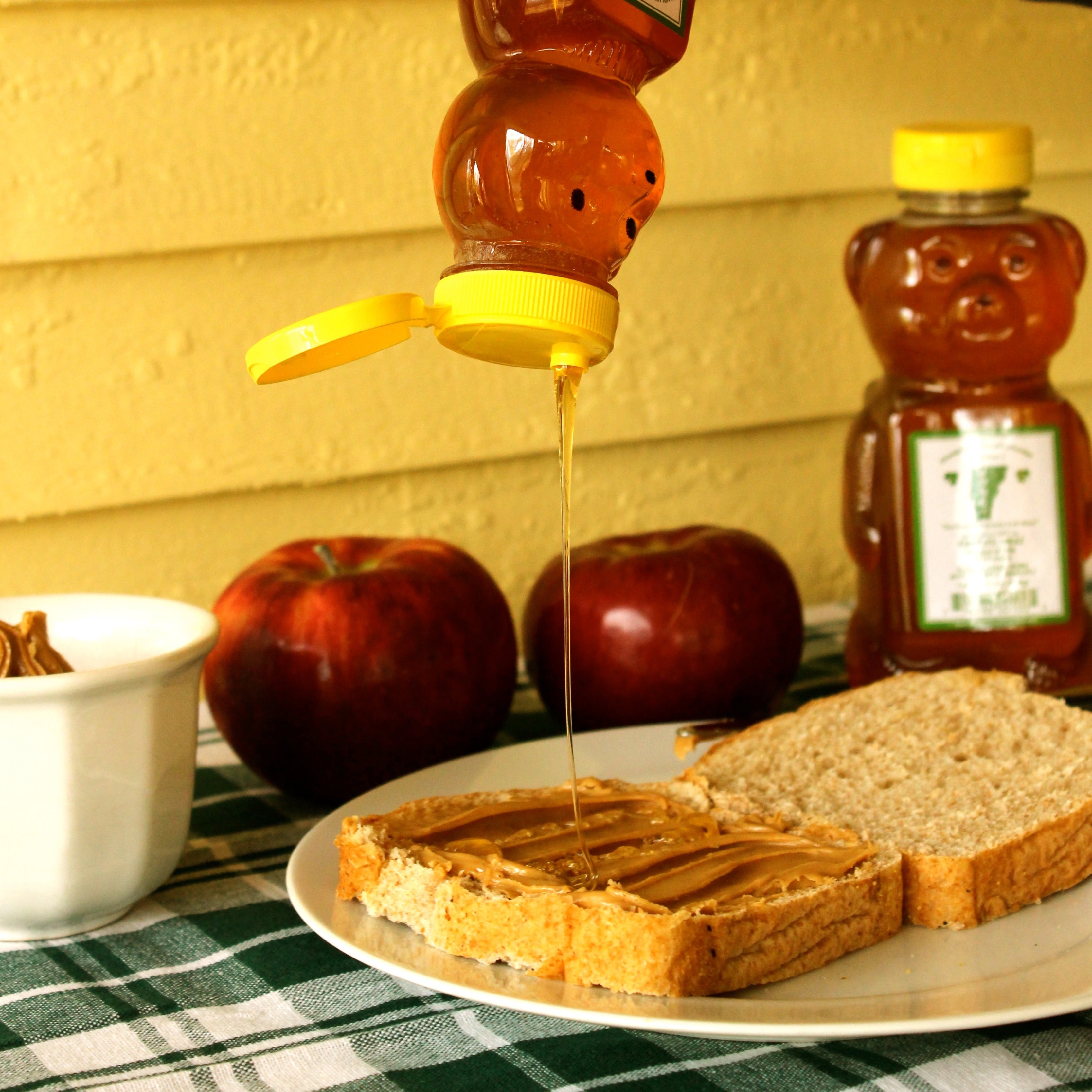 Honey being poured from a honey bear onto an open peanut butter sandwich on a plate with apples and in the background